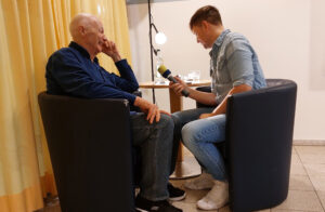 N-Joy Reporter interviewt einen älteren Mann in einem Sessel, beide sitzen einander gegenüber.
