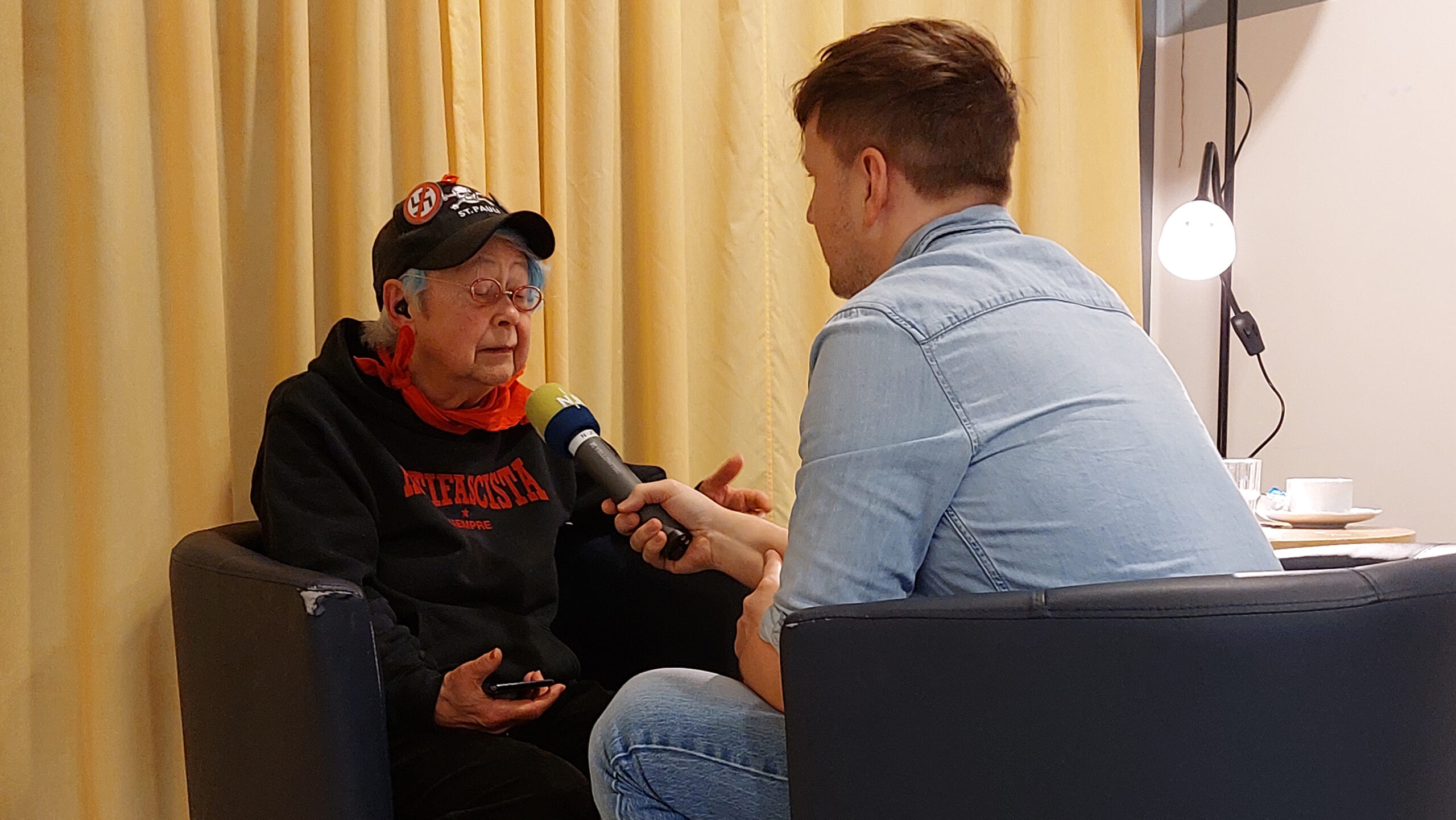Ein Journalist interviewt eine ältere Frau in einem Sessel, beide sitzen einander gegenüber.