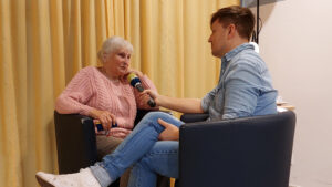 Ein Journalist interviewt eine ältere Frau in einem Sessel, beide sitzen einander gegenüber.